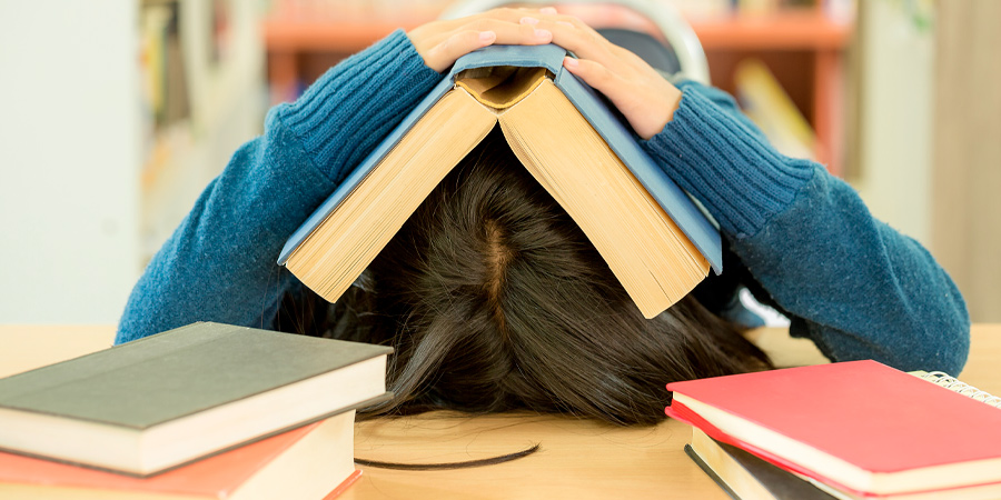Aluno com a cabeça sobre a mesa e um livro aberto sobre a cabeça